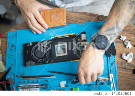 Person repairs a graphics card on a blue work surface with various tools and parts around in a home setting during daytime Person repairs a graphics card on a blue work surface with various tools and parts around in a home setting during daytime 141023601