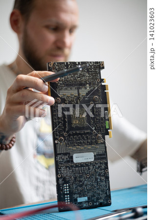 Man works on a computer graphics card while holding it in one hand and using tools with the other hand in a well-lit room Man works on a computer graphics card while holding it in one hand and using tools with the other hand in a well-lit room 141023603