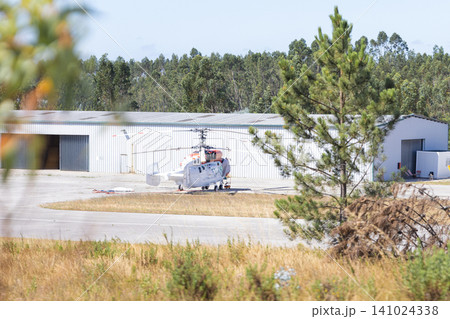 Helicopter parked on a tarmac near a large hangar surrounded by trees and grass, showcasing a clear blue sky in the background 141024338