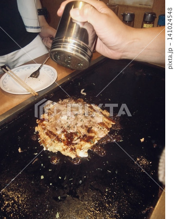 鉄板の上で焼き上がる熱々のお好み焼きと調味料を振る手元 鉄板の上で焼き上がる熱々のお好み焼きと調味料を振る手元 141024548