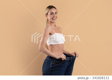Smiling young woman in oversized jeans posing sideways, showcasing dramatic weight loss Smiling young woman in oversized jeans posing sideways, showcasing dramatic weight loss 141026111