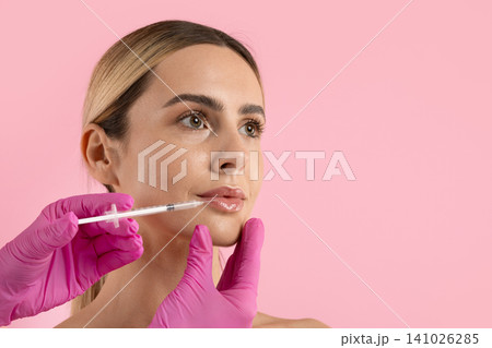 Young woman receiving cosmetic lip filler injection with syringe on pink background 141026285