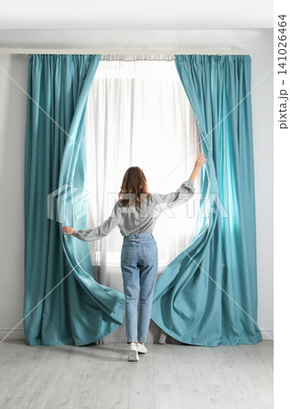 Woman opening window curtains at home in morning, back view Woman opening window curtains at home in morning, back view 141026464