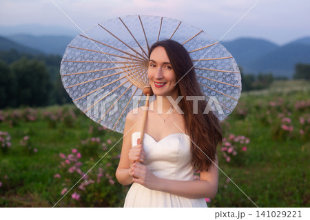 Elegant young woman in white dress holding paper umbrella in rose field Elegant young woman in white dress holding paper umbrella in rose field 141029221