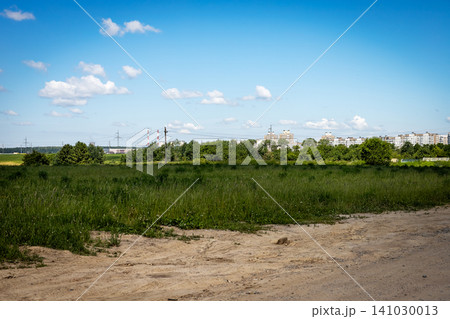 Open grassy field with dirt path, mixed green vegetation, wildflowers, distant apartment buildings, bright blue sky and scattered clouds on clear summer horizon 141030013