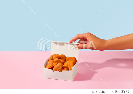 Hand opens takeout box filled with crispy chicken nuggets on pink and blue background. Hand opens takeout box filled with crispy chicken nuggets on pink and blue background. 141030577