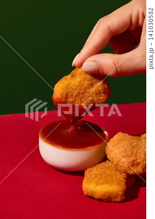 Hand dips crispy chicken nugget into ketchup sauce on red and green background Hand dips crispy chicken nugget into ketchup sauce on red and green background 141030582