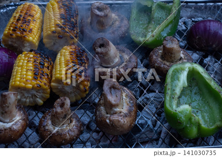 Close-up of various vegetables including corn on the cob, mushrooms and green bell peppers grilling on a wire rack with smoke rising from hot charcoal 141030735