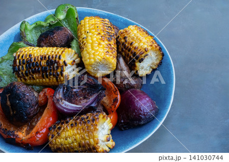 Grilled vegetables including corn on the cob, mushrooms, red onion and green bell peppers served on a blue plate over a grey background with copy space 141030744