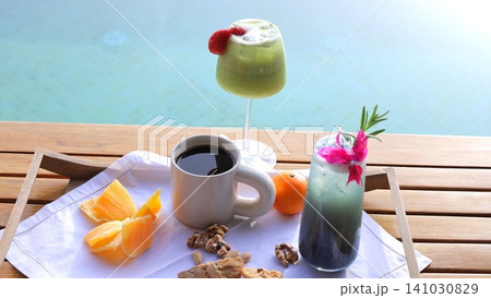 Poolside breakfast arrangement at 5 star hotel with coffee, green smoothie, iced cocktail, fresh fruit, and tropical garnish on sunlit wooden deck beside turquoise water and pool 141030829