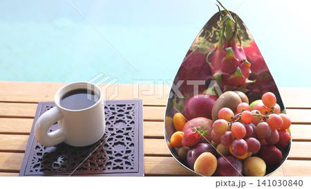 Breakfast coffee beside polished metal bowl of fresh tropical fruit on wooden table near pool in bright resort setting with colorful grapes, strawberries, plums, kumquats, dragon fruit, apricots 141030840