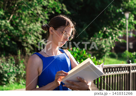 Woman enjoying reading book in green park. Intentional technologies break. Slow living lifestyle expressed in warm day reading and cognitive reset from digital overload Woman enjoying reading book in green park. Intentional technologies break. Slow living lifestyle expressed in warm day reading and cognitive reset from digital overload 141031266