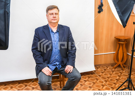A man sits on a chair in a photo studio, preparing for a photo shoot for documents and a passport A man sits on a chair in a photo studio, preparing for a photo shoot for documents and a passport 141031381