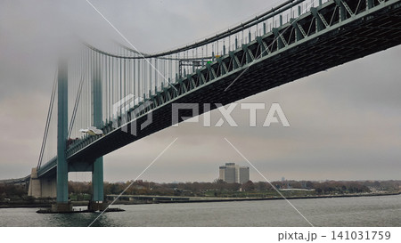 Horizontal low angle view of a massive suspension bridge steel structure disappearing into thick white fog with a single seagull bird flying near the river bank Horizontal low angle view of a massive suspension bridge steel structure disappearing into thick white fog with a single seagull bird flying near the river bank 141031759