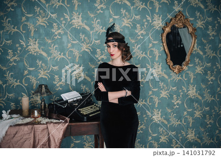 Elegant girl in 1920s black velvet dress posing with arms crossed by vintage typewriter Elegant girl in 1920s black velvet dress posing with arms crossed by vintage typewriter 141031792