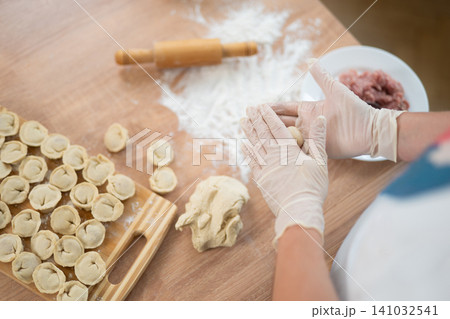 Woman in gloves shaping dough for handmade dumplings on a floured wooden table Woman in gloves shaping dough for handmade dumplings on a floured wooden table 141032541
