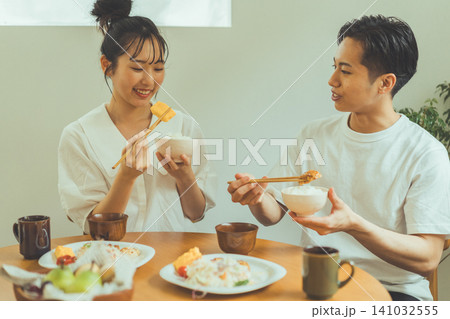ダイニングテーブルで玉子焼きとご飯を食べながら笑顔で会話するカップルの食事シーン ダイニングテーブルで玉子焼きとご飯を食べながら笑顔で会話するカップルの食事シーン 141032555