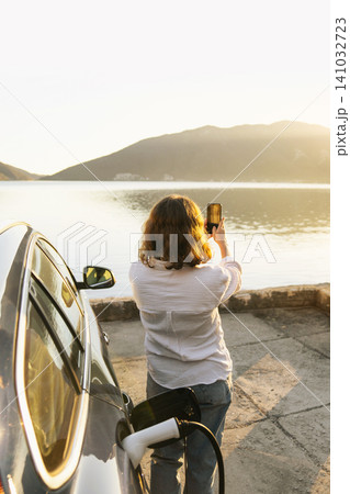 Woman with smartphone next to charging electric car on the seacost during summer vacation. Woman with smartphone next to charging electric car on the seacost during summer vacation. 141032723