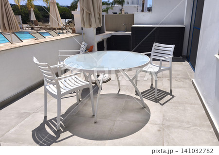 Modern outdoor patio with white table and chairs beside a swimming pool, featuring a sunny terrace and resort atmosphere, ideal for relaxing, dining, and enjoying a summer vacation 141032782