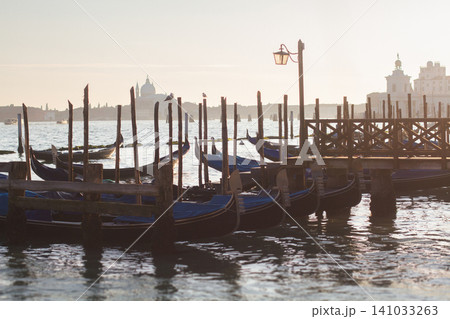 Traditional Venetian gondolas moored at a wooden pier in the lagoon during a hazy sunrise in Venice Traditional Venetian gondolas moored at a wooden pier in the lagoon during a hazy sunrise in Venice 141033263