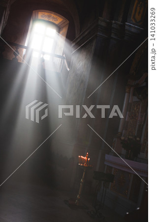 Atmospheric sunlight streaming through high church window illuminating dark interior with burning candles in cathedral Atmospheric sunlight streaming through high church window illuminating dark interior with burning candles in cathedral 141033269