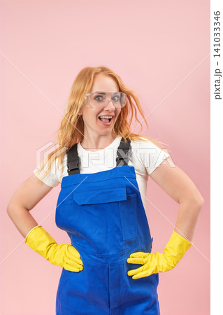 Cheerful red haired woman with safety goggles and yellow gloves standing confidently with hands on hips, expressing excitement and readiness for work against a pink background Cheerful red haired woman with safety goggles and yellow gloves standing confidently with hands on hips, expressing excitement and readiness for work against a pink background 141033346