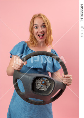 Excited woman holding a worn vehicle steering wheel, expressing surprise and readiness for a new journey, standing against a pink studio background 141033364