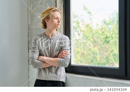 Thoughtful teenage boy with crossed arms by window Thoughtful teenage boy with crossed arms by window 141034104