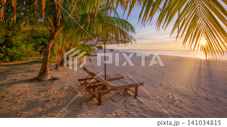 Warm dusk light bathing palm beach sun loungers. Tropical shore glowing under a golden evening sky. Romantic island retreat for luxury travel summer holiday Warm dusk light bathing palm beach sun loungers. Tropical shore glowing under a golden evening sky. Romantic island retreat for luxury travel summer holiday 141034815