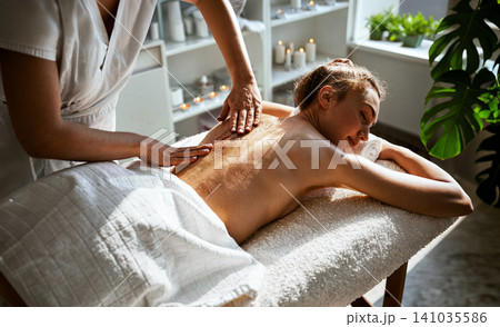 Golden Body Scrub Treatment Therapist Massages Glittering Exfoliant into Womans Back on White Towel Table With Candles and Lush Plants in Spa Room Golden Body Scrub Treatment Therapist Massages Glittering Exfoliant into Womans Back on White Towel Table With Candles and Lush Plants in Spa Room 141035586