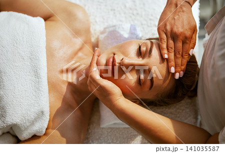 Deep Relaxation Facial Massage Close Up Therapist Hands Cradle and Press Glowing Womans Face as She Rests Eyes Closed on Bright White Spa Table Deep Relaxation Facial Massage Close Up Therapist Hands Cradle and Press Glowing Womans Face as She Rests Eyes Closed on Bright White Spa Table 141035587