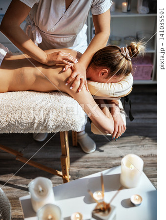 Candlelit Spa Close Up of Therapist Hands Applying Pressure to Womans Upper Back and Shoulder Blade on Soft Plush Massage Table With Candles in 141035591