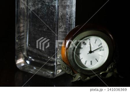 Table clock and glass bottle on dark background Table clock and glass bottle on dark background 141036369