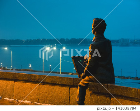 Volgograd, Russia  July 5, 2023 Sculpture. A man reads a book while sitting on the embankment parapet at dusk, set against the backdrop of city lights 141038794