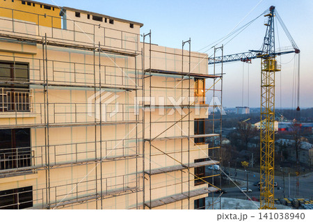 Apartment or office tall unfinished building under construction. Brick wall in scaffolding, shiny windows and tower crane on urban landscape and blue sky background. Drone aerial photography. Apartment or office tall unfinished building under construction. Brick wall in scaffolding, shiny windows and tower crane on urban landscape and blue sky background. Drone aerial photography. 141038940