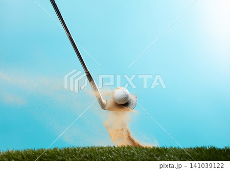Golf ball explode from Tee off. Golfer hit ball with metal iron club to sand explosion on green. Golf club hit ball on green tee off with sand flying scatter. Blue Sky background isolated Golf ball explode from Tee off. Golfer hit ball with metal iron club to sand explosion on green. Golf club hit ball on green tee off with sand flying scatter. Blue Sky background isolated 141039122
