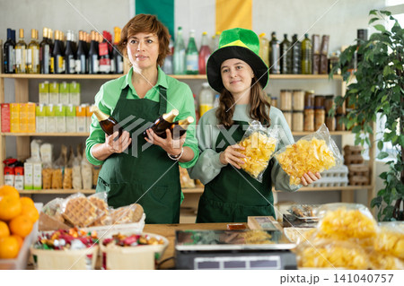 Two female sellers posing in green costume and hat, offering bottle of irish ale in store decorated for St. Patrick Day Two female sellers posing in green costume and hat, offering bottle of irish ale in store decorated for St. Patrick Day 141040757