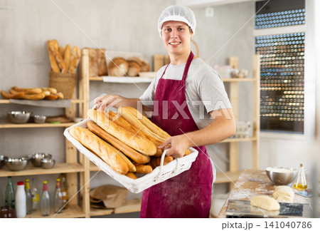 European young bakery male seller stands proudly in front of fresh bread holding basket of freshly baked baguettes in kitchen European young bakery male seller stands proudly in front of fresh bread holding basket of freshly baked baguettes in kitchen 141040786