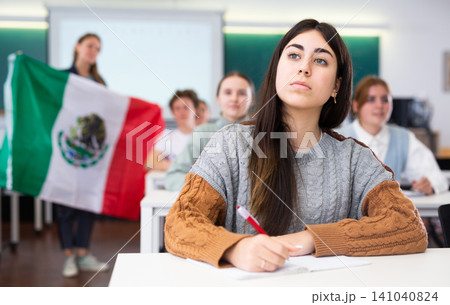 Female teacher tells her classmates about the country of Mexico, holding flag in her hands Female teacher tells her classmates about the country of Mexico, holding flag in her hands 141040824