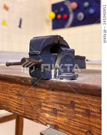 Blue bench vise tightened on a sturdy wooden workbench in a workshop classroom, symbolizing hands on vocational training, woodworking, trades skills and practical education Blue bench vise tightened on a sturdy wooden workbench in a workshop classroom, symbolizing hands on vocational training, woodworking, trades skills and practical education 141040951