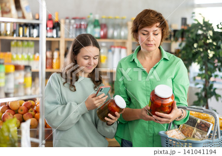 Woman with her daughter choose pickled tomatoes with a phone in hands Woman with her daughter choose pickled tomatoes with a phone in hands 141041194