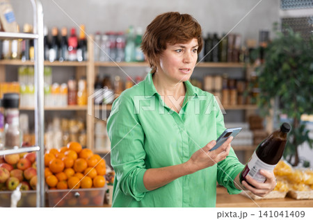 European woman customer at shop scans QR code on pomegranate juice bottle using phone camera European woman customer at shop scans QR code on pomegranate juice bottle using phone camera 141041409