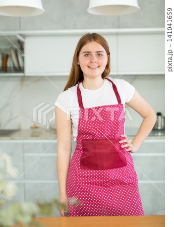 Positive teen girl wearing red polka dot apron smiling at camera while preparing for cooking in kitchen Positive teen girl wearing red polka dot apron smiling at camera while preparing for cooking in kitchen 141041659