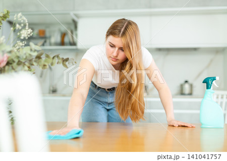 Attentive European teen girl in casual clothes wiping dust on countertop with microfiber rag while cleaning cozy cuisine Attentive European teen girl in casual clothes wiping dust on countertop with microfiber rag while cleaning cozy cuisine 141041757
