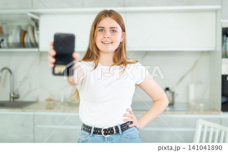 Portrait of positive teen girl showing modern mobile phone with black screen in cozy kitchen. Gadget with empty free space for mock up Portrait of positive teen girl showing modern mobile phone with black screen in cozy kitchen. Gadget with empty free space for mock up 141041890