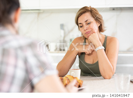 Upset middle-aged woman talking sadly while drinking tea in the kitchen Upset middle-aged woman talking sadly while drinking tea in the kitchen 141041947