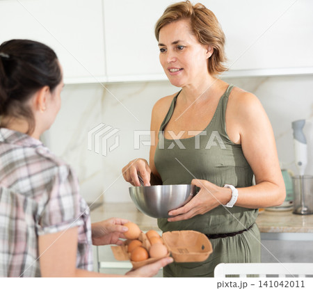 Middle-aged woman holding bowl talking to another woman in the kitchen 141042011