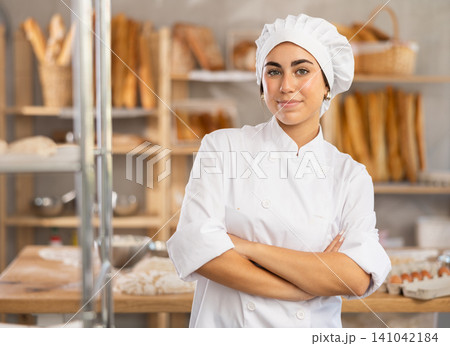 Young female baker stands in bake house, ready to greet customers Young female baker stands in bake house, ready to greet customers 141042184