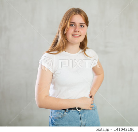 Portrait of cheerful charismatic teen girl in white t-shirt and blue jeans posing against gray wall with copy space Portrait of cheerful charismatic teen girl in white t-shirt and blue jeans posing against gray wall with copy space 141042186