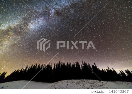 Winter mountains night landscape panorama. Milky Way bright constellation in dark blue starry sky over dark spruce pine trees forest, soft glow on horizon after sunset. Wide angle shot. 141043867